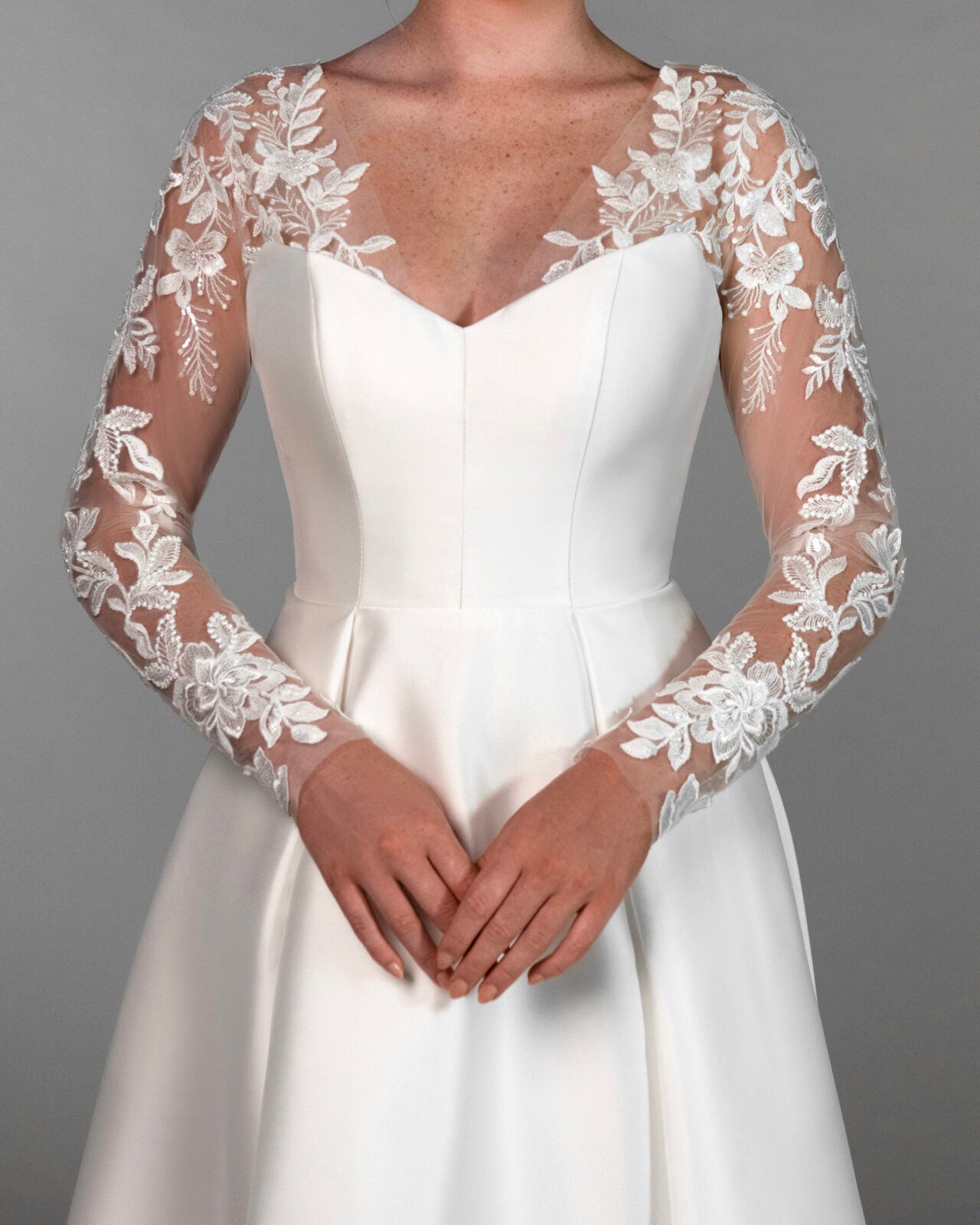 A woman wearing a white wedding dress with long sheer lace sleeves and a fitted bodice. Lace Stella York jacket sleeves over satin dress