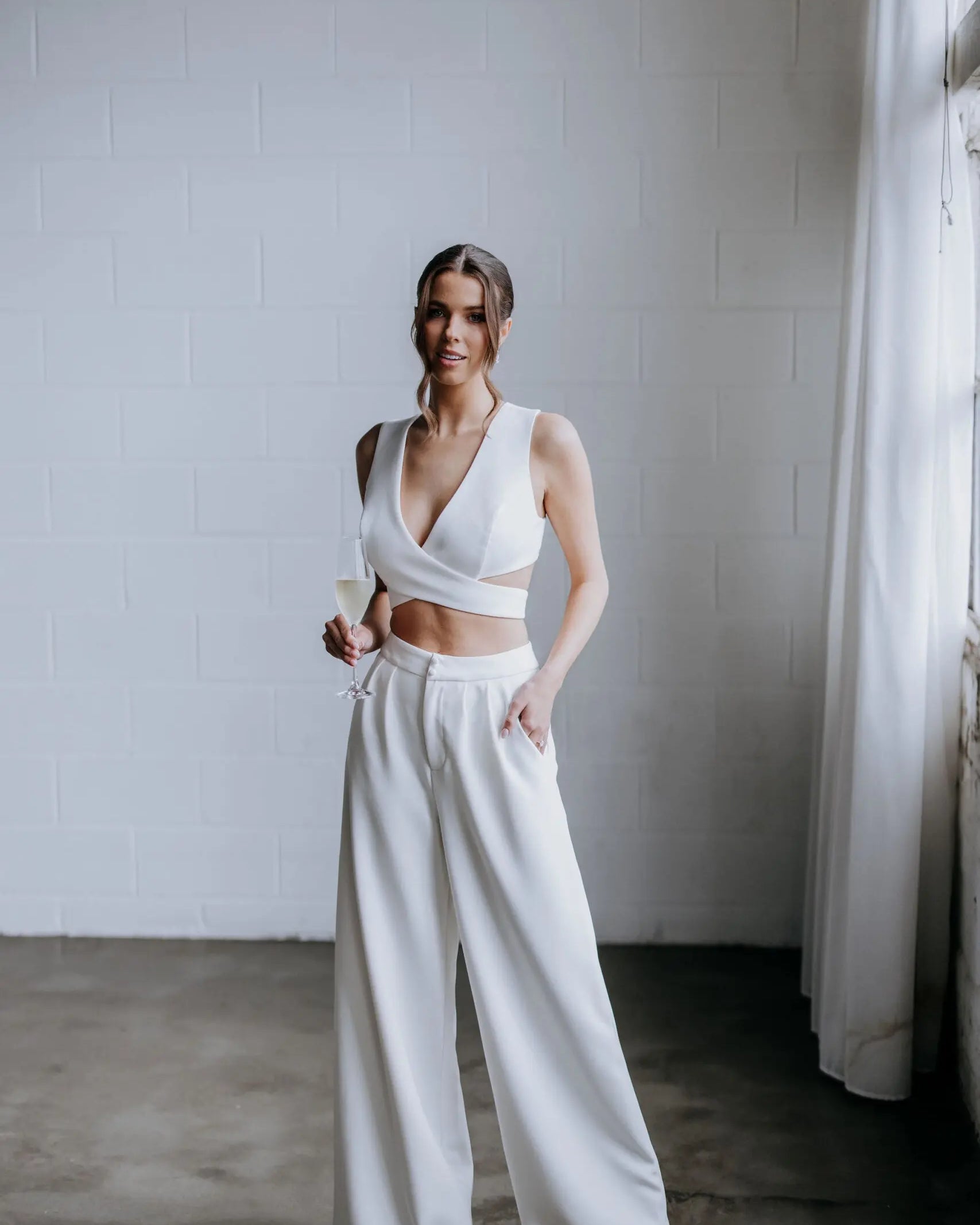 Model wearing the Hailey two-piece pant suit by Kellylin Couture in ivory. High-waisted tailored pants paired with a criss-cross wrap-around crop top. Model facing forward, hand in pocket, holding a glass of champagne. Elegant bridal-inspired look.
