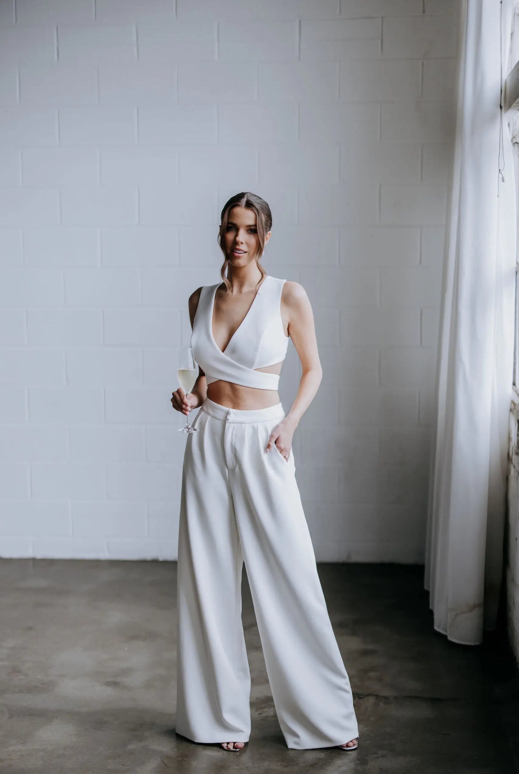 Model wearing the Hailey two-piece pant suit by Kellylin Couture in ivory. High-waisted tailored pants paired with a criss-cross wrap-around crop top. Model facing forward, hand in pocket, holding a glass of champagne. Elegant bridal-inspired look.