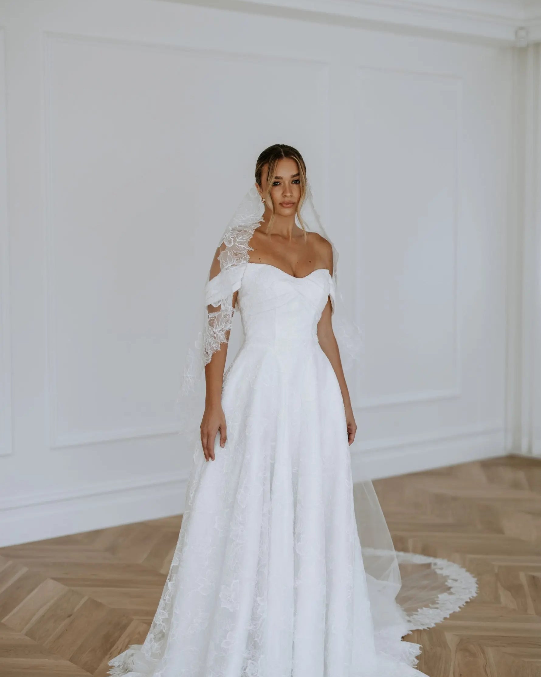 A bride wearing an off-the-shoulder lace A-line wedding dress with a sweetheart neckline, floral lace detailing, and a long train — elegant modern bridal gown photographed in a bright studio.