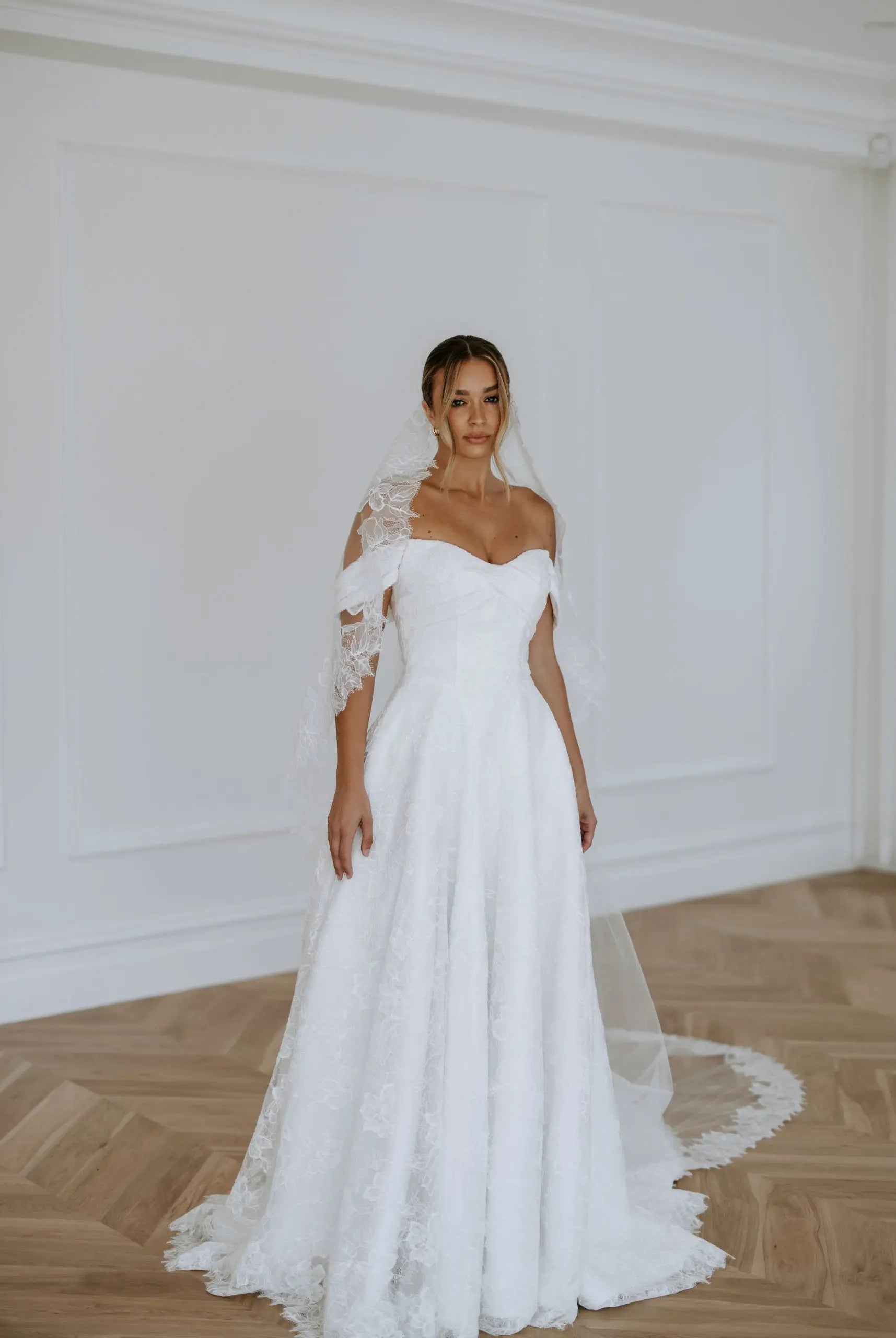 A bride wearing an off-the-shoulder lace A-line wedding dress with a sweetheart neckline, floral lace detailing, and a long train — elegant modern bridal gown photographed in a bright studio.