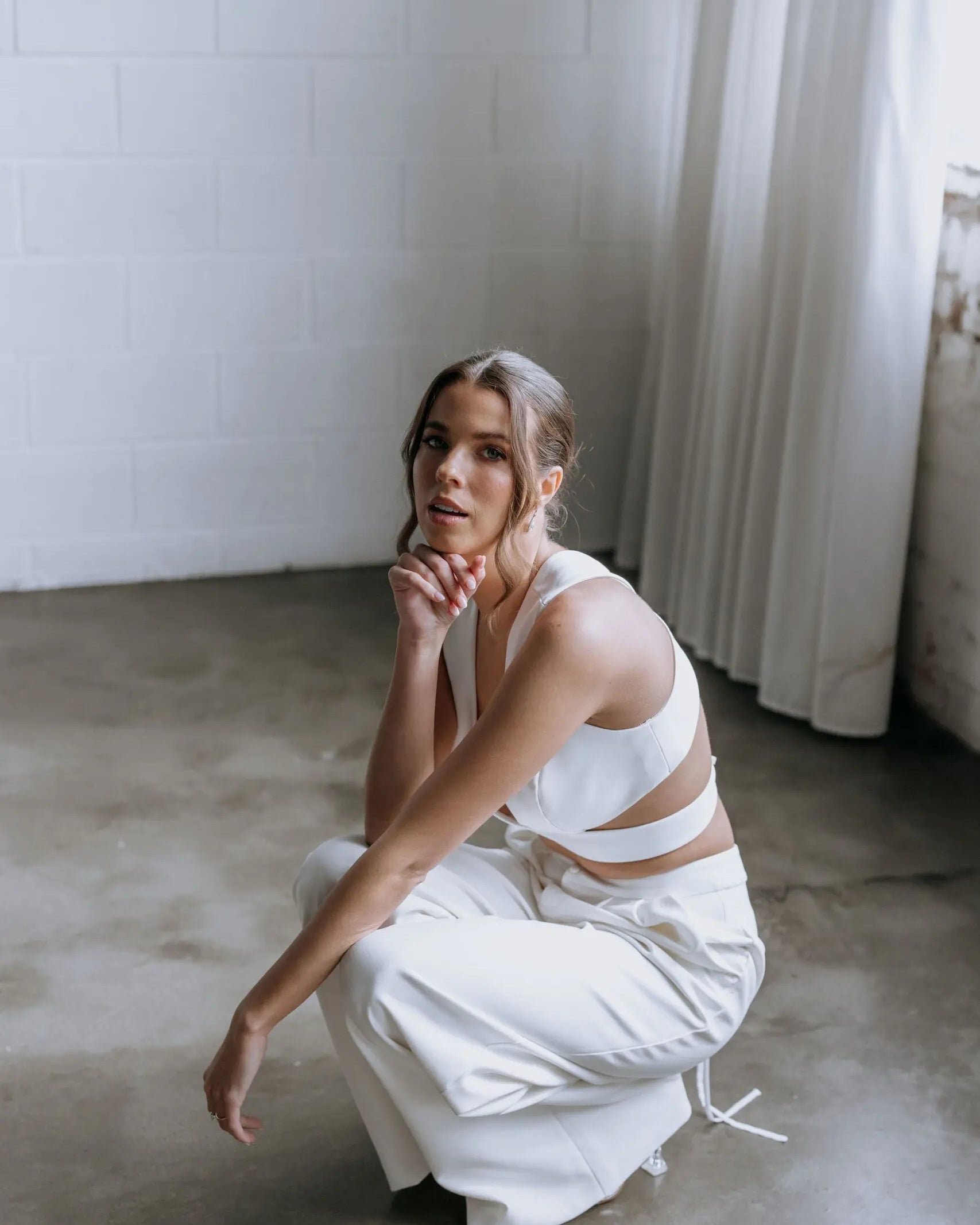 Model kneeling while wearing the Hailey two-piece pant suit by Kellylin Couture in ivory. High-waisted tailored pants paired with a criss-cross wrap-around crop top, showcasing the stylish back detail. Elegant bridal pant suit perfect for showers, engagement parties, or rehearsal dinners.