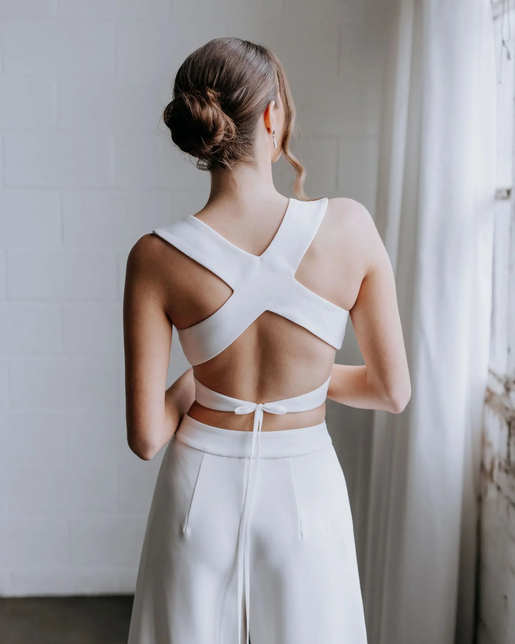 Model wearing the Hailey two-piece pant suit by Kellylin Couture in ivory. High-waisted tailored pants paired with a criss-cross wrap-around crop top with a stylish back detail. Model facing forward, hand in pocket, holding a glass of champagne. Elegant bridal pant suit perfect for showers, engagement parties, or rehearsal dinners.