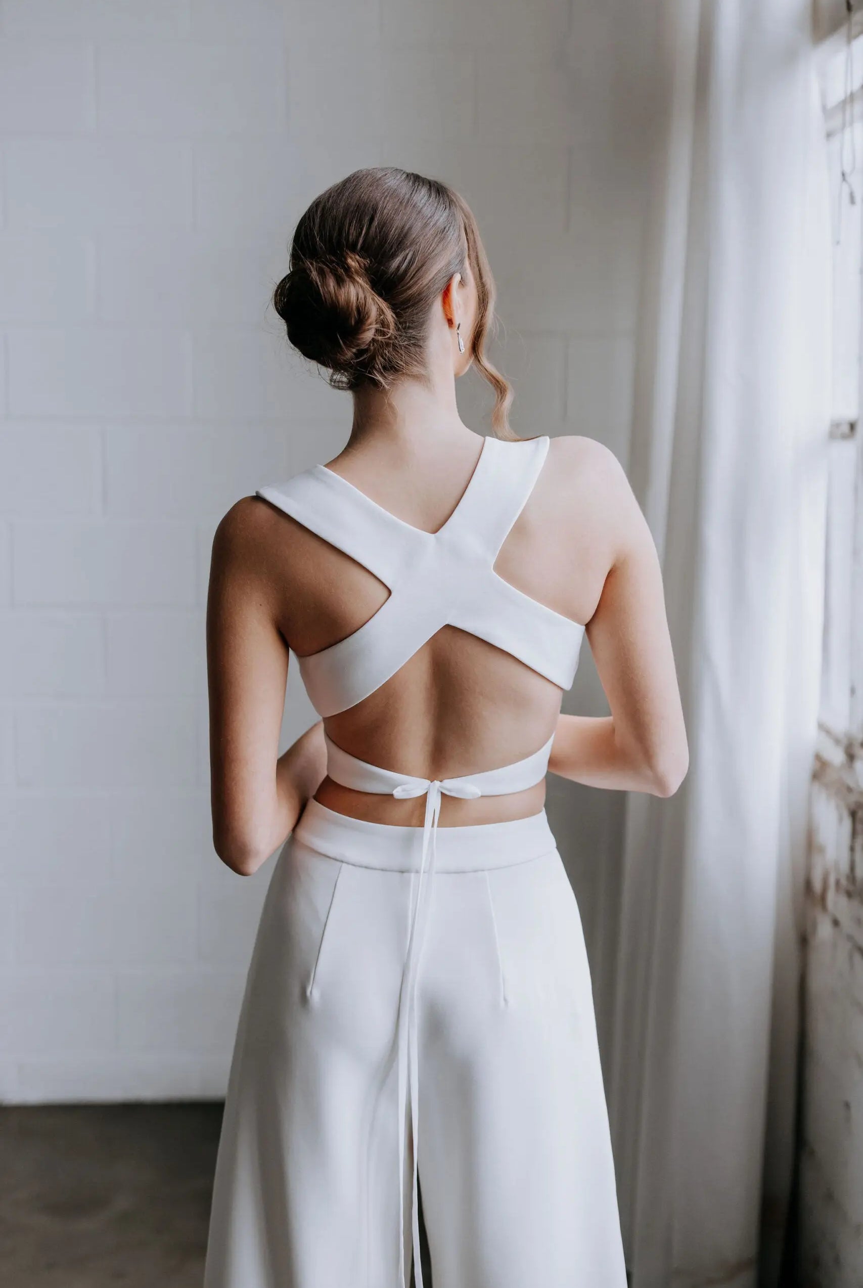 Model wearing the Hailey two-piece pant suit by Kellylin Couture in ivory. High-waisted tailored pants paired with a criss-cross wrap-around crop top with a stylish back detail. Model facing forward, hand in pocket, holding a glass of champagne. Elegant bridal pant suit perfect for showers, engagement parties, or rehearsal dinners.
