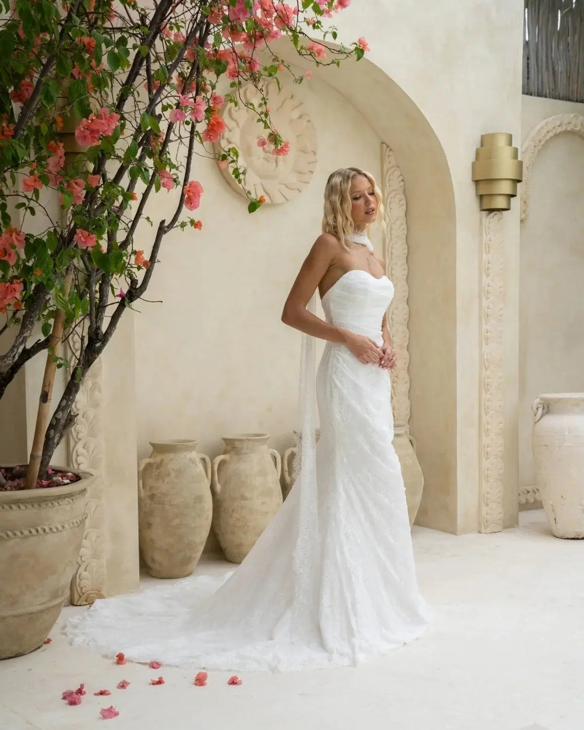 A bride in a strapless white lace wedding dress stands in a soft, Mediterranean-style setting, surrounded by pink flowers and clay pots. She poses calmly, showing the gown’s sleek, straight silhouette and gentle draping.