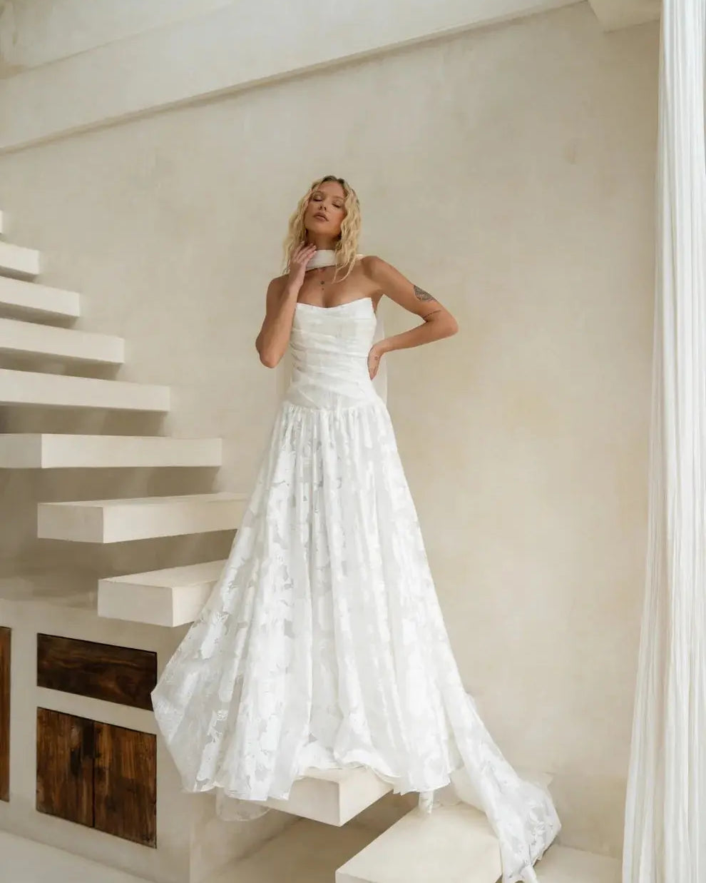 A bride wearing a strapless floral Jacquard A-line wedding gown poses on modern floating stairs in a soft, minimalist interior, styled with the optional Tonica Scarf.