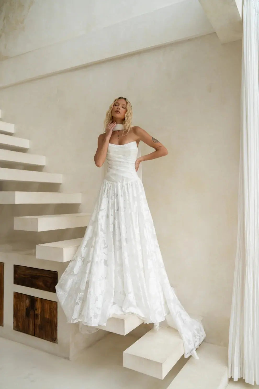 A bride wearing a strapless floral Jacquard A-line wedding gown poses on modern floating stairs in a soft, minimalist interior, styled with the optional Tonica Scarf.