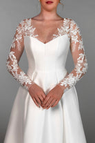 A woman wearing a white wedding dress with long sheer lace sleeves and a fitted bodice. Lace Stella York jacket sleeves over satin dress