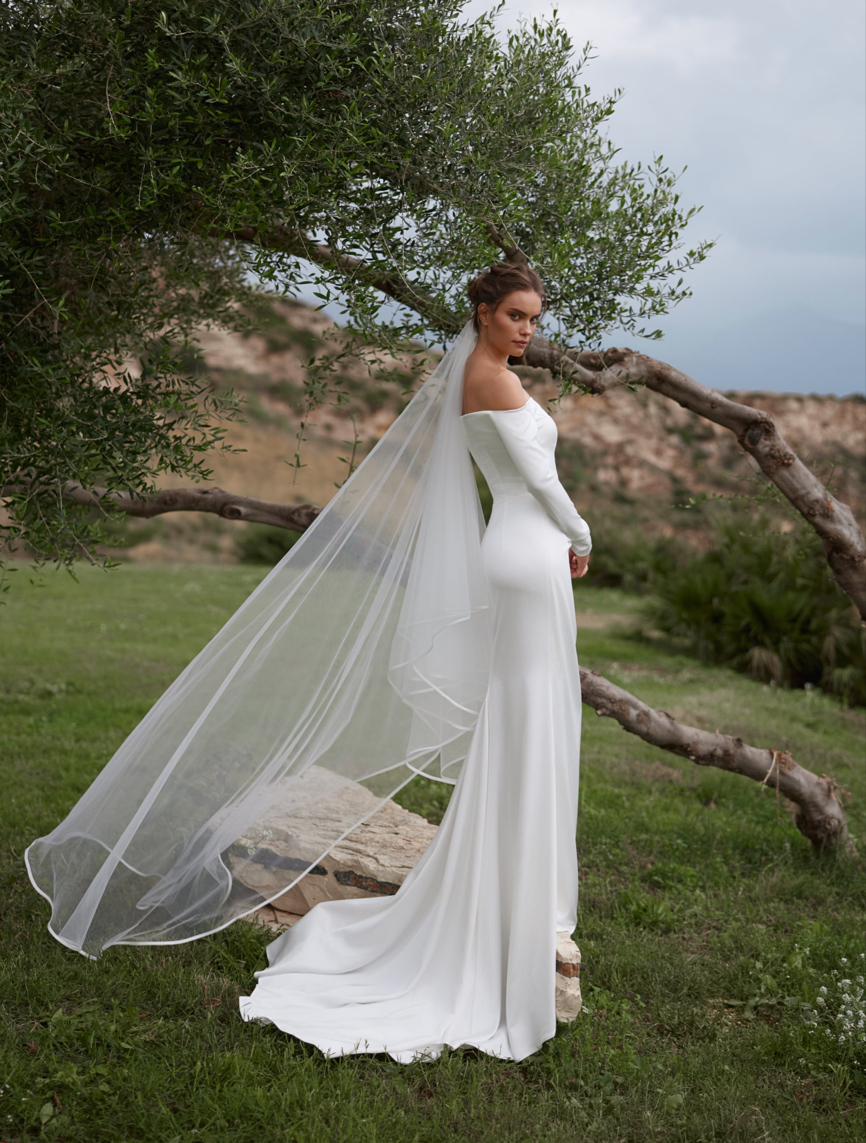 A woman wearing a long sleeve off-the-shoulder white wedding dress with a flowing sheer veil.