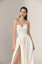 A woman modeling a strapless white bridal gown with a sweetheart neckline, ruched bodice, high front slit, and flowing skirt.