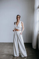 Model wearing the Hailey two-piece pant suit by Kellylin Couture in ivory. High-waisted tailored pants paired with a criss-cross wrap-around crop top. Model facing forward, hand in pocket, holding a glass of champagne. Elegant bridal-inspired look.