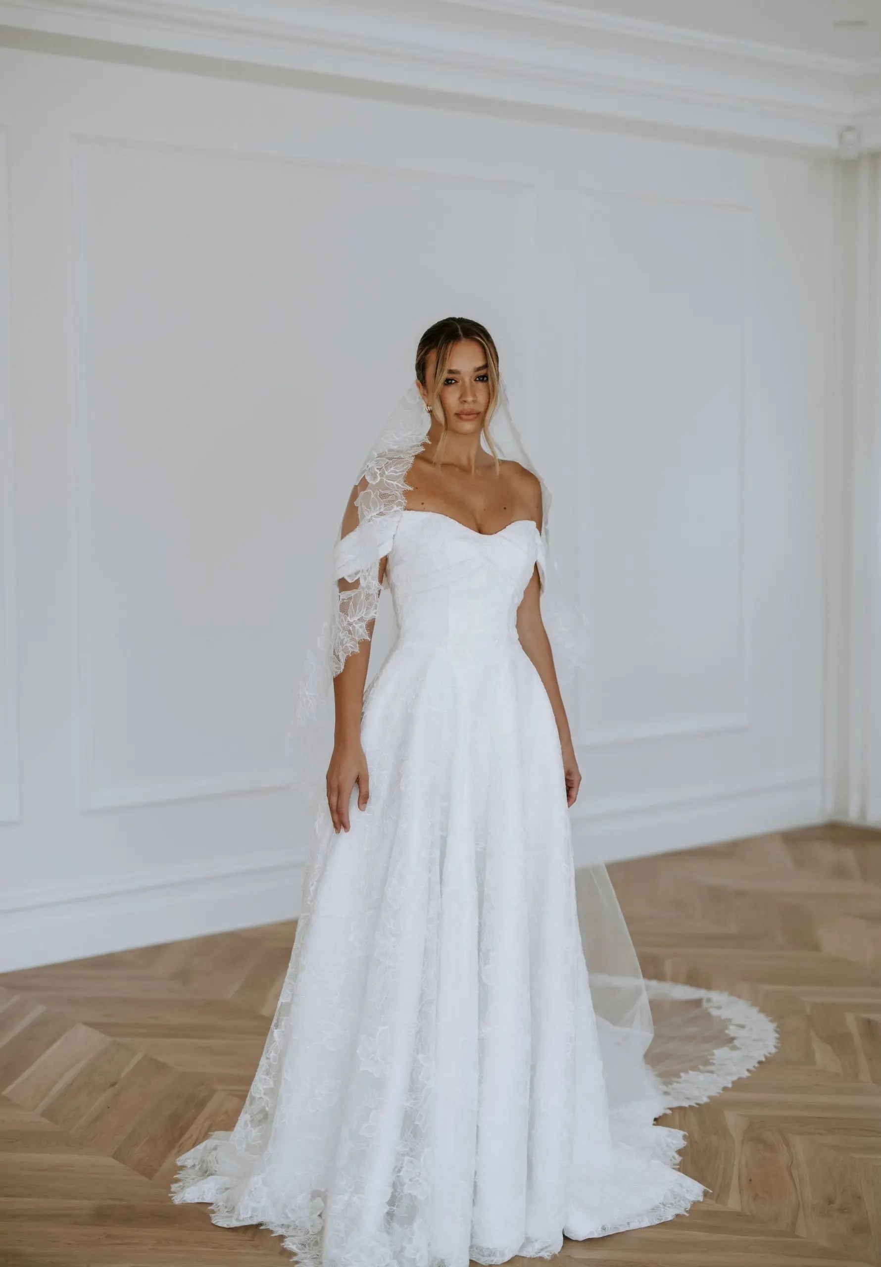 A bride wearing an off-the-shoulder lace A-line wedding dress with a sweetheart neckline, floral lace detailing, and a long train — elegant modern bridal gown photographed in a bright studio.