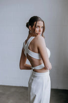 Model looking over her shoulder wearing the Hailey two-piece pant suit by Kellylin Couture in ivory. High-waisted tailored pants paired with a criss-cross wrap-around crop top, showcasing the stylish back detail. Elegant bridal pant suit for showers, engagement parties, or rehearsal dinners.