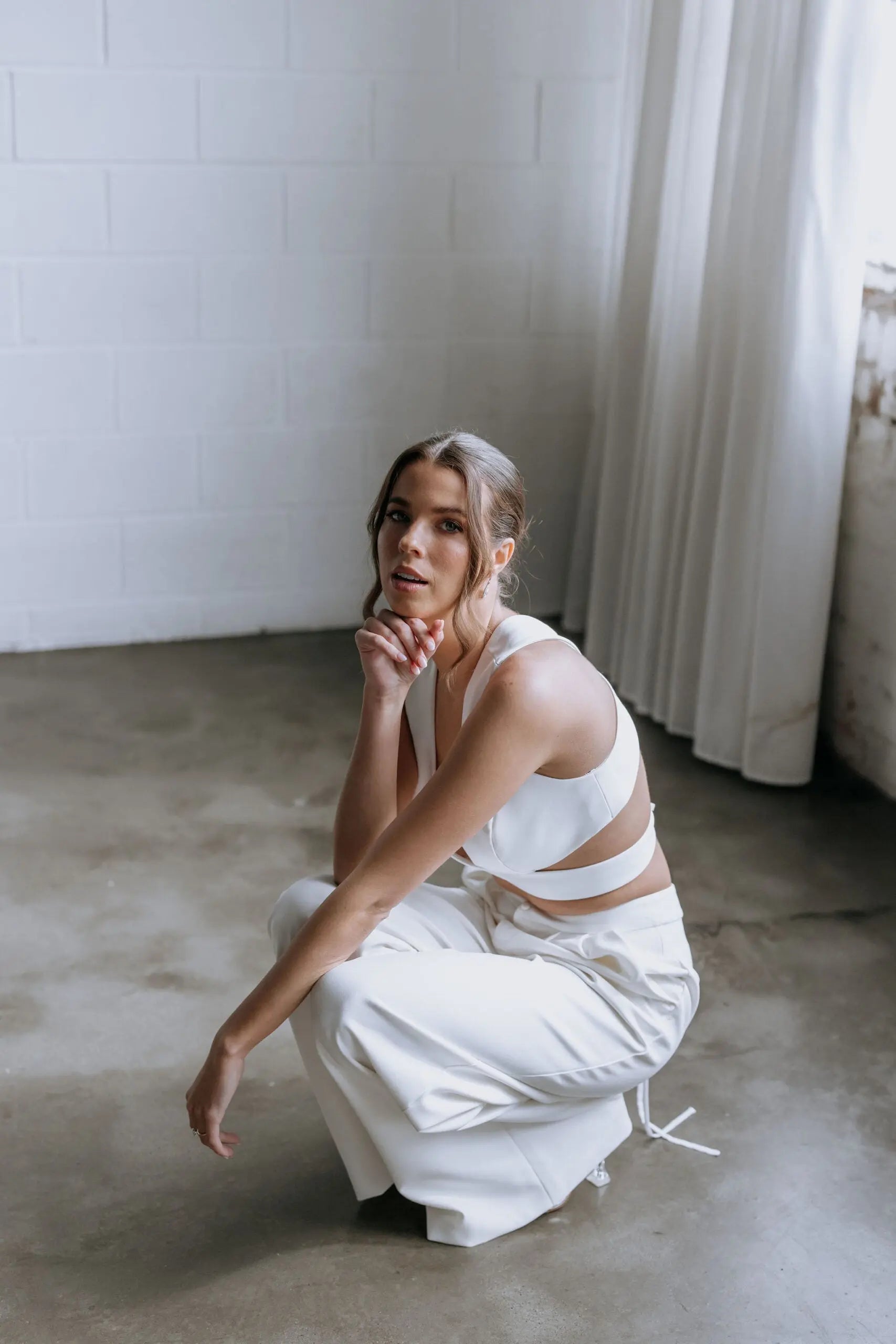 Model kneeling while wearing the Hailey two-piece pant suit by Kellylin Couture in ivory. High-waisted tailored pants paired with a criss-cross wrap-around crop top, showcasing the stylish back detail. Elegant bridal pant suit perfect for showers, engagement parties, or rehearsal dinners.