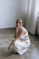 Model kneeling while wearing the Hailey two-piece pant suit by Kellylin Couture in ivory. High-waisted tailored pants paired with a criss-cross wrap-around crop top, showcasing the stylish back detail. Elegant bridal pant suit perfect for showers, engagement parties, or rehearsal dinners.