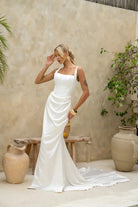 A bride wearing the Katara wedding gown by Jessica Couture stands in a soft, neutral-toned courtyard. The gown features a square neckline, thin straps, and gentle draping at the waist.