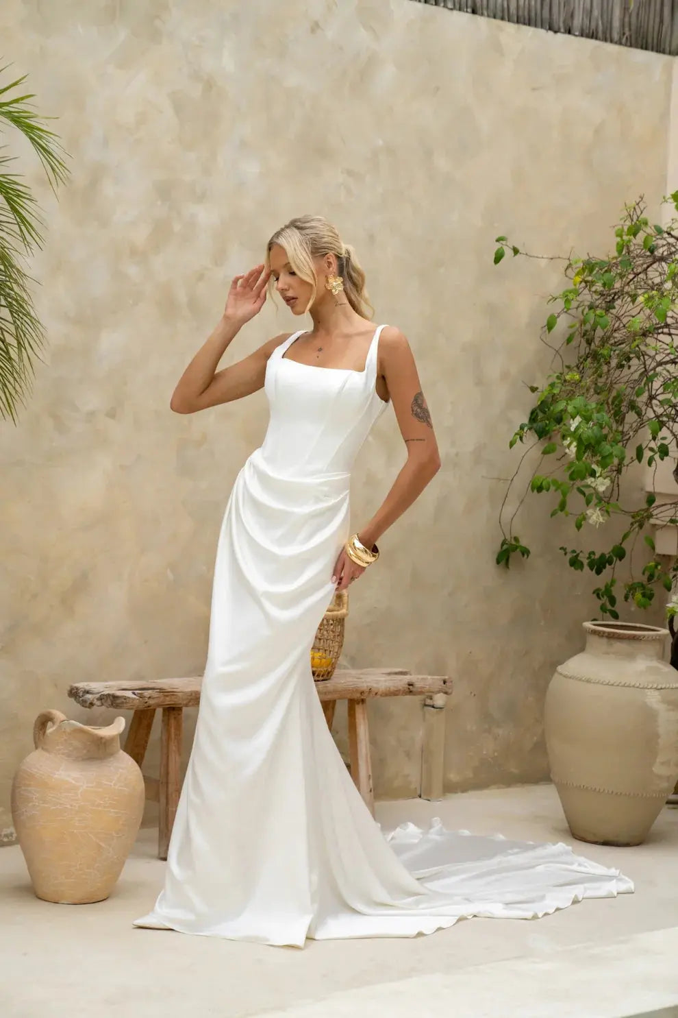 A bride wearing the Katara wedding gown by Jessica Couture stands in a soft, neutral-toned courtyard. The gown features a square neckline, thin straps, and gentle draping at the waist.