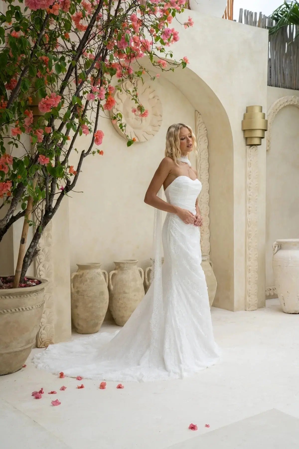 A bride in a strapless white lace wedding dress stands in a soft, Mediterranean-style setting, surrounded by pink flowers and clay pots. She poses calmly, showing the gown’s sleek, straight silhouette and gentle draping.